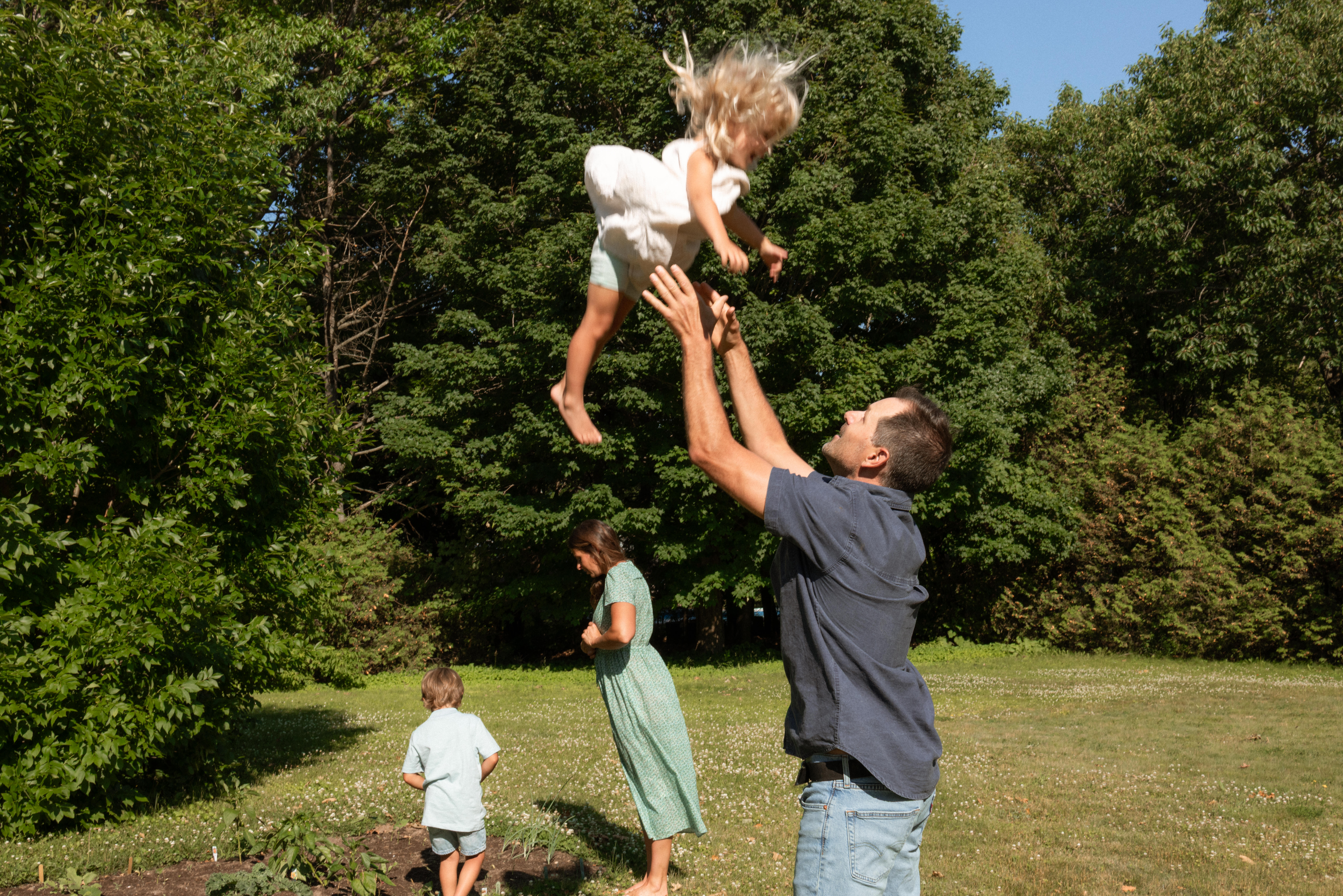 candid family moment in documentary family session. mother exploring the garden with her son as dad throws daughter up in the air 