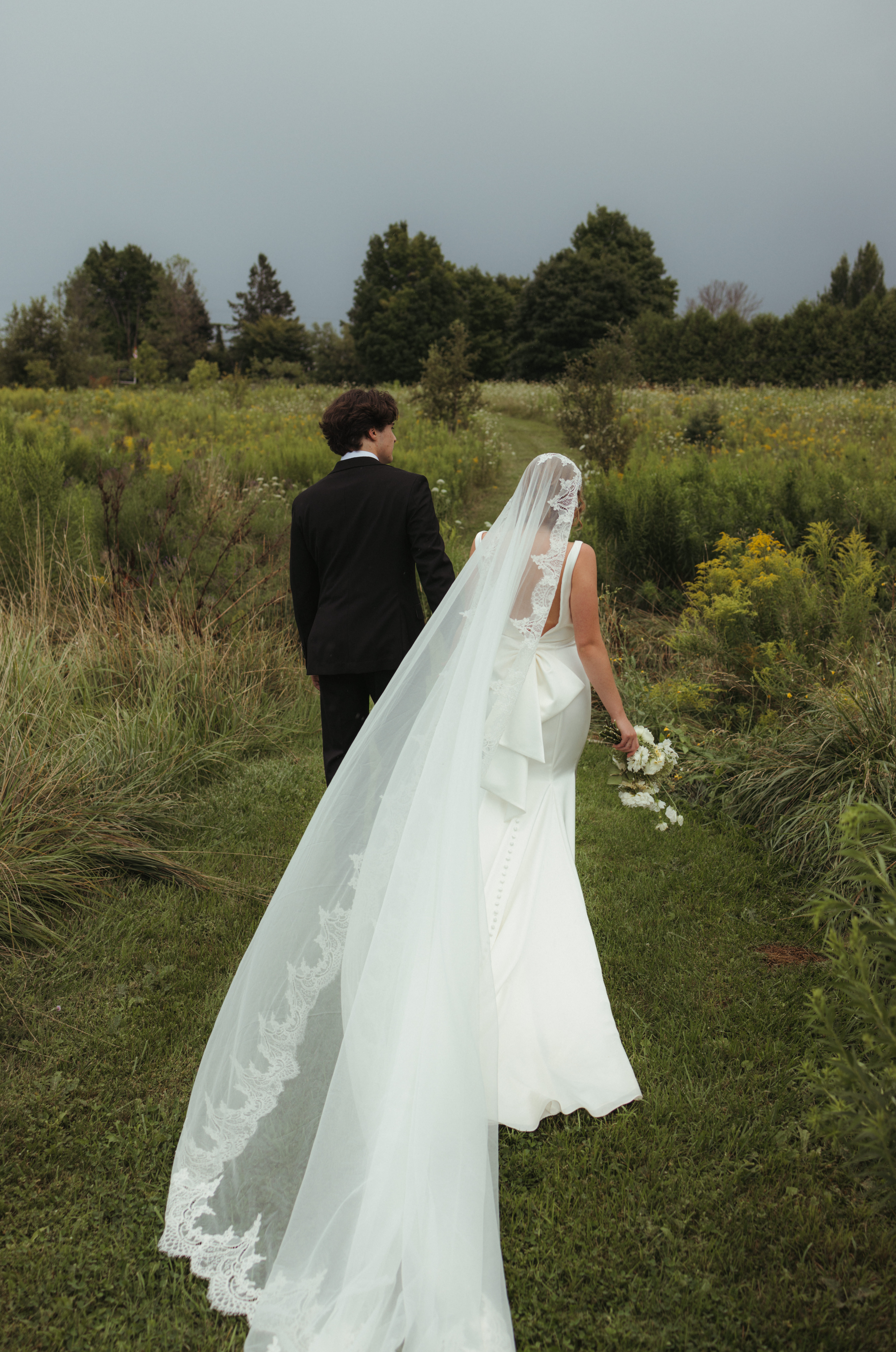 bride and groom walking in open field as the brides veil blows in the wind